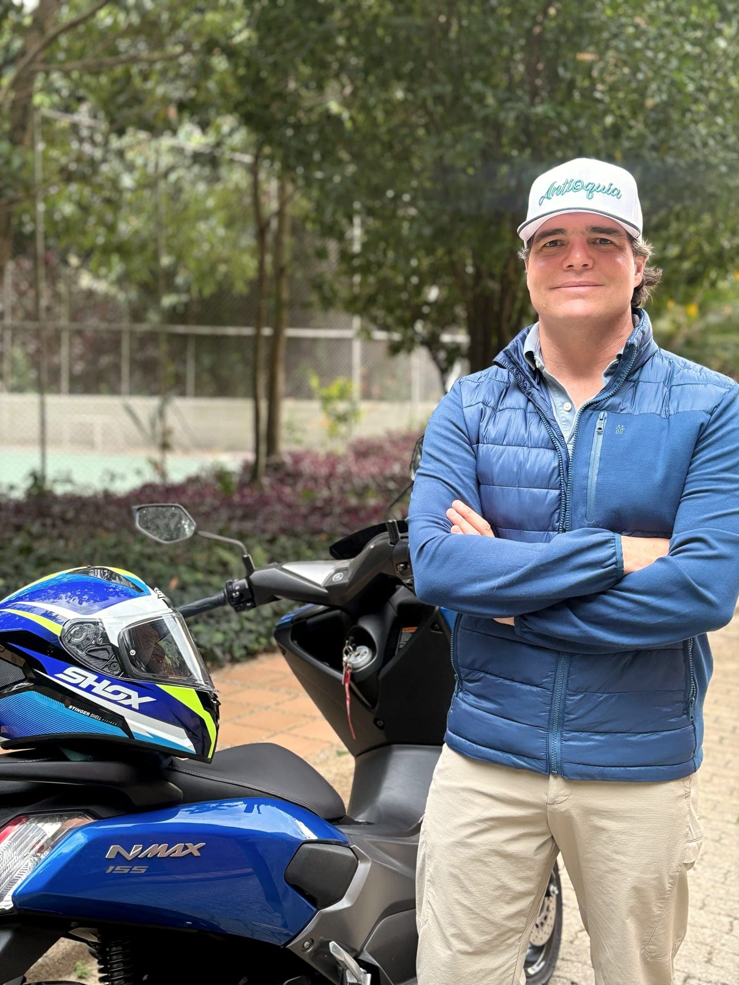Alejandro Arbeláez A. de brazos cruzados junto a su moto, con chaqueta azul y gorra de Antioquia, bajo los árboles