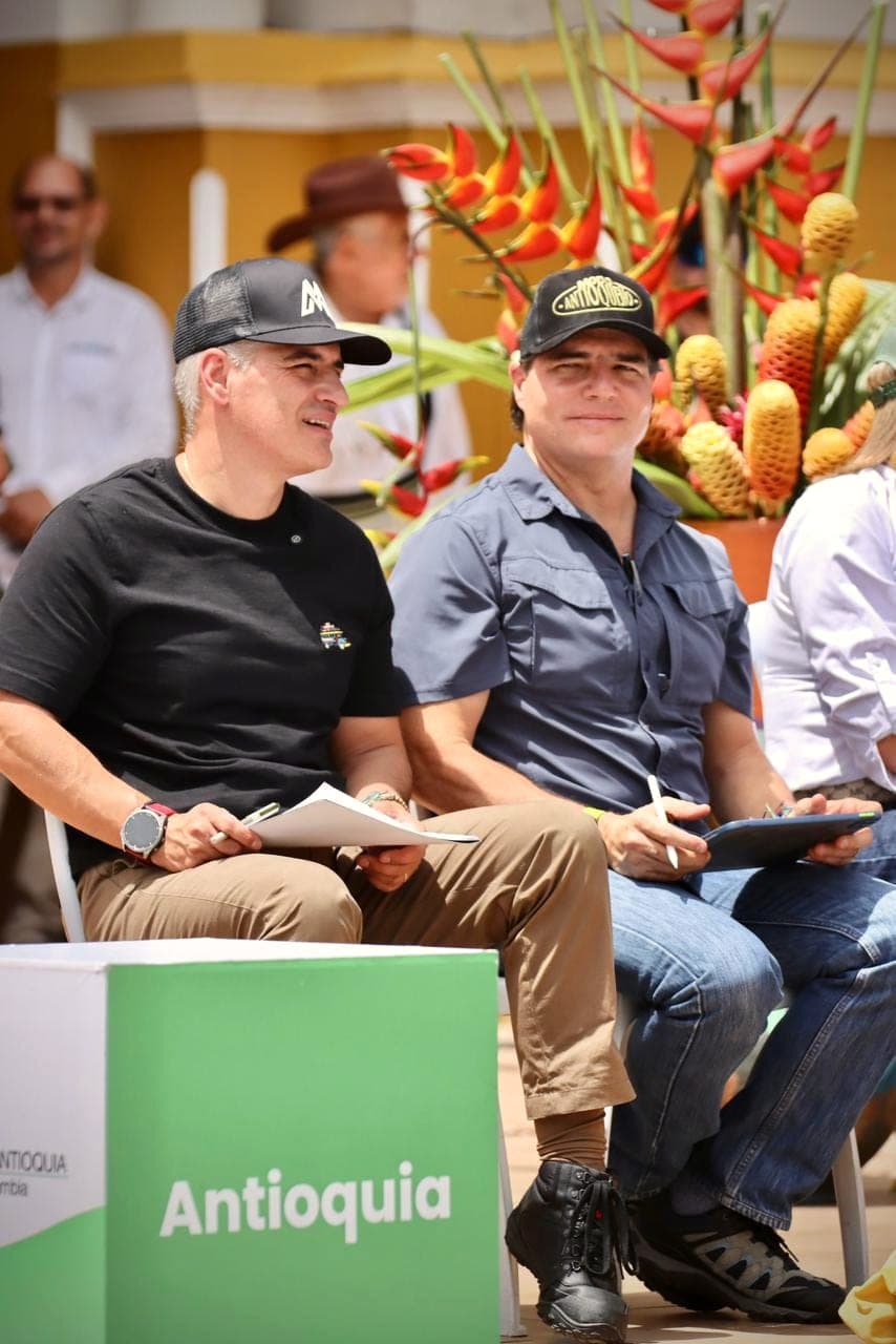 Alejandro Arbeláez A. y el gobernador Andrés Julián Rendón, sonrientes y con gorra, sentados en un acto al aire libre con flores de heliconia detrás y una caja con la palabra Antioquia