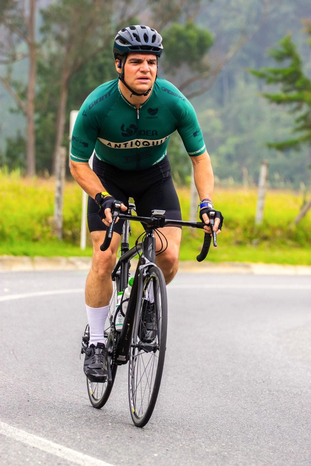 Alejandro Arbeláez A. sube una montaña en bicicleta de ruta, con camiseta verde y casco, en una carretera entre pastizales